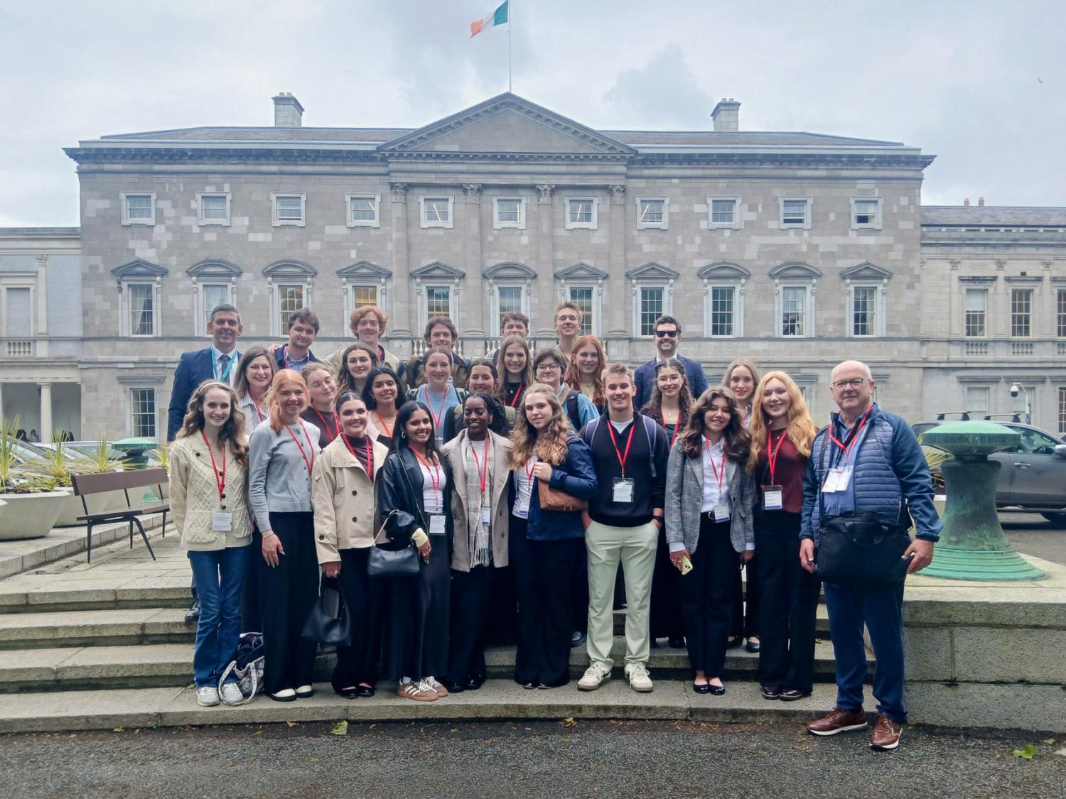 Group of adults posing in front of a historic government building with an Irish flag overhead on a cloudy day.