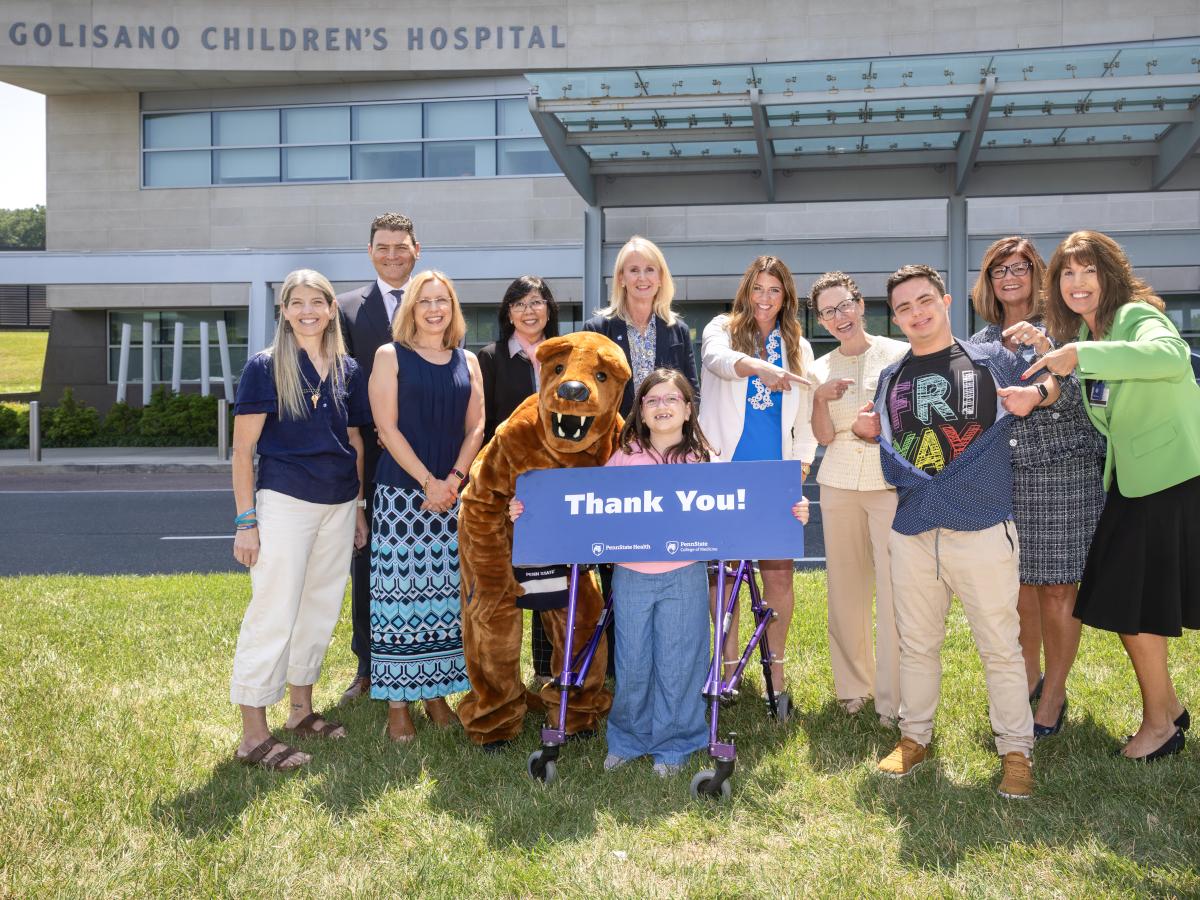 A group of people and a costumed Nittany Lion pose for a photo in front of Penn State Health Golisano Children’s Hospital, many of them smiling and pointing, “goofy pose” style. They hold a large sign that reads “Thank You!”