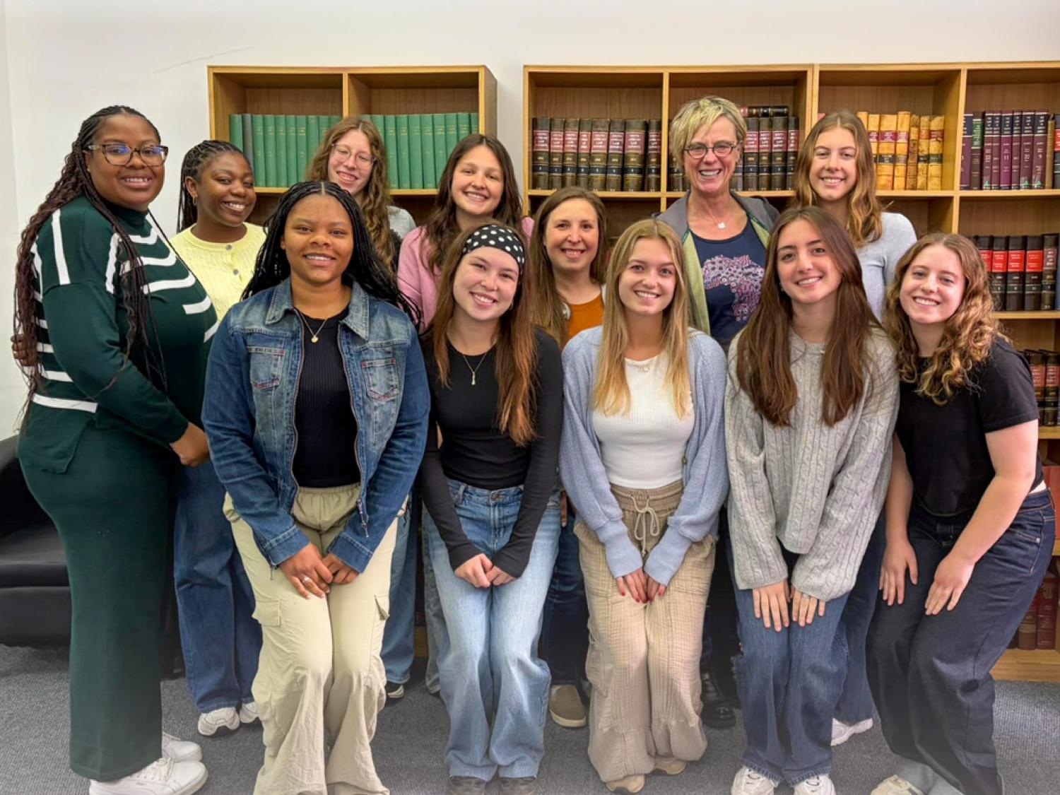 A group of 13 individuals poses for a photo in a library setting, smiling and dressed in casual clothing. Behind them are wooden shelves filled with books.