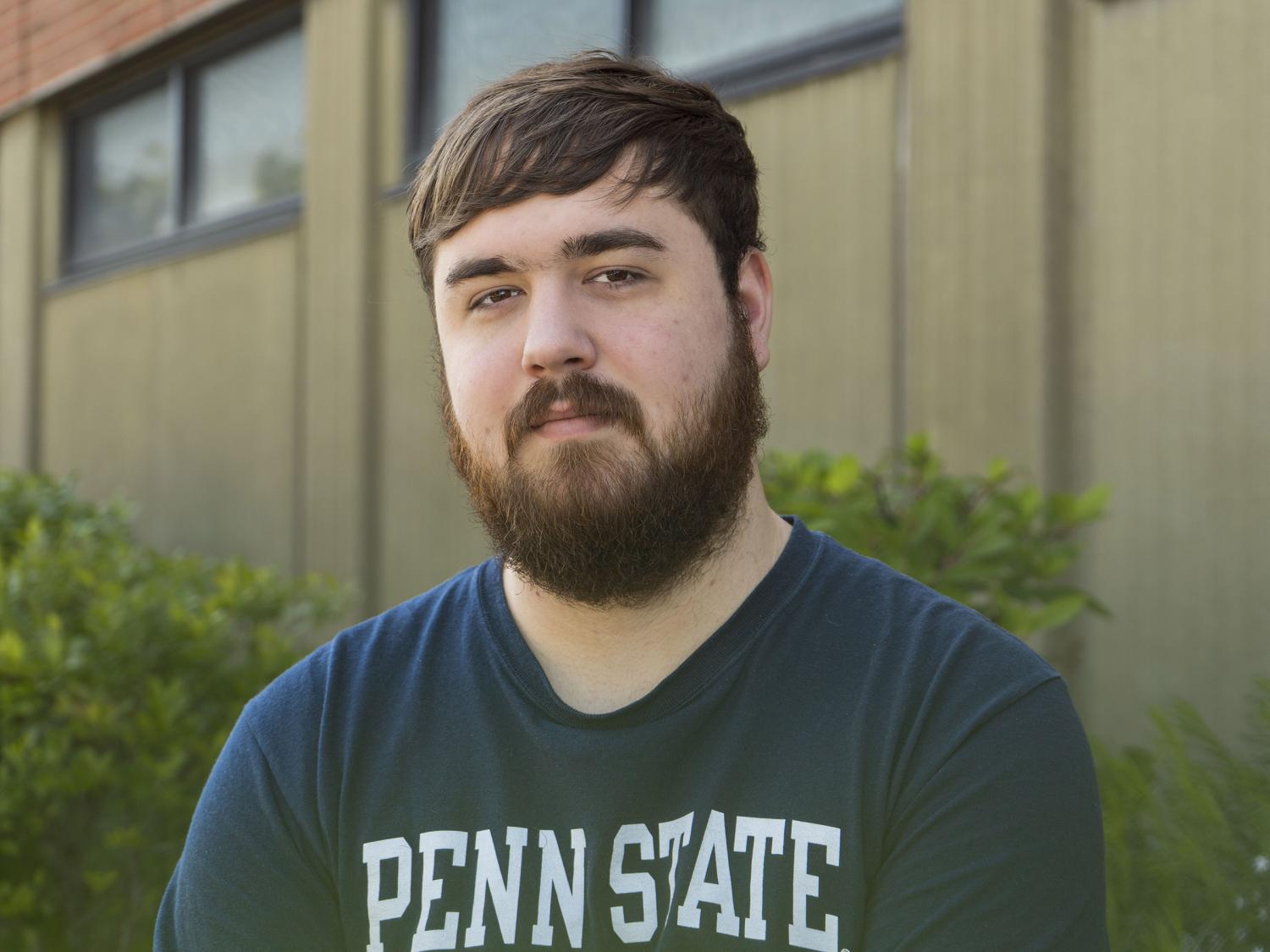 Male student wearing navy Penn State shirt smiles with brick building in background.