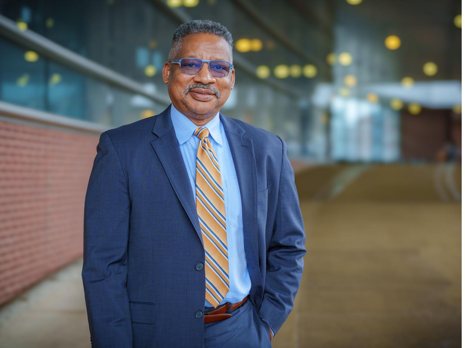 person with gray mustache and glasses in blue business suit with brick and glass building in background 