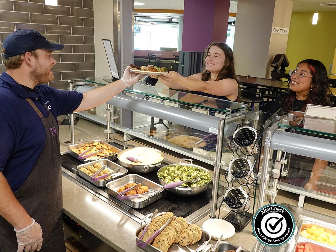 A staff member at Pure Dining Station in the East Food District serves food to two female students (AllerCheck Seal included)