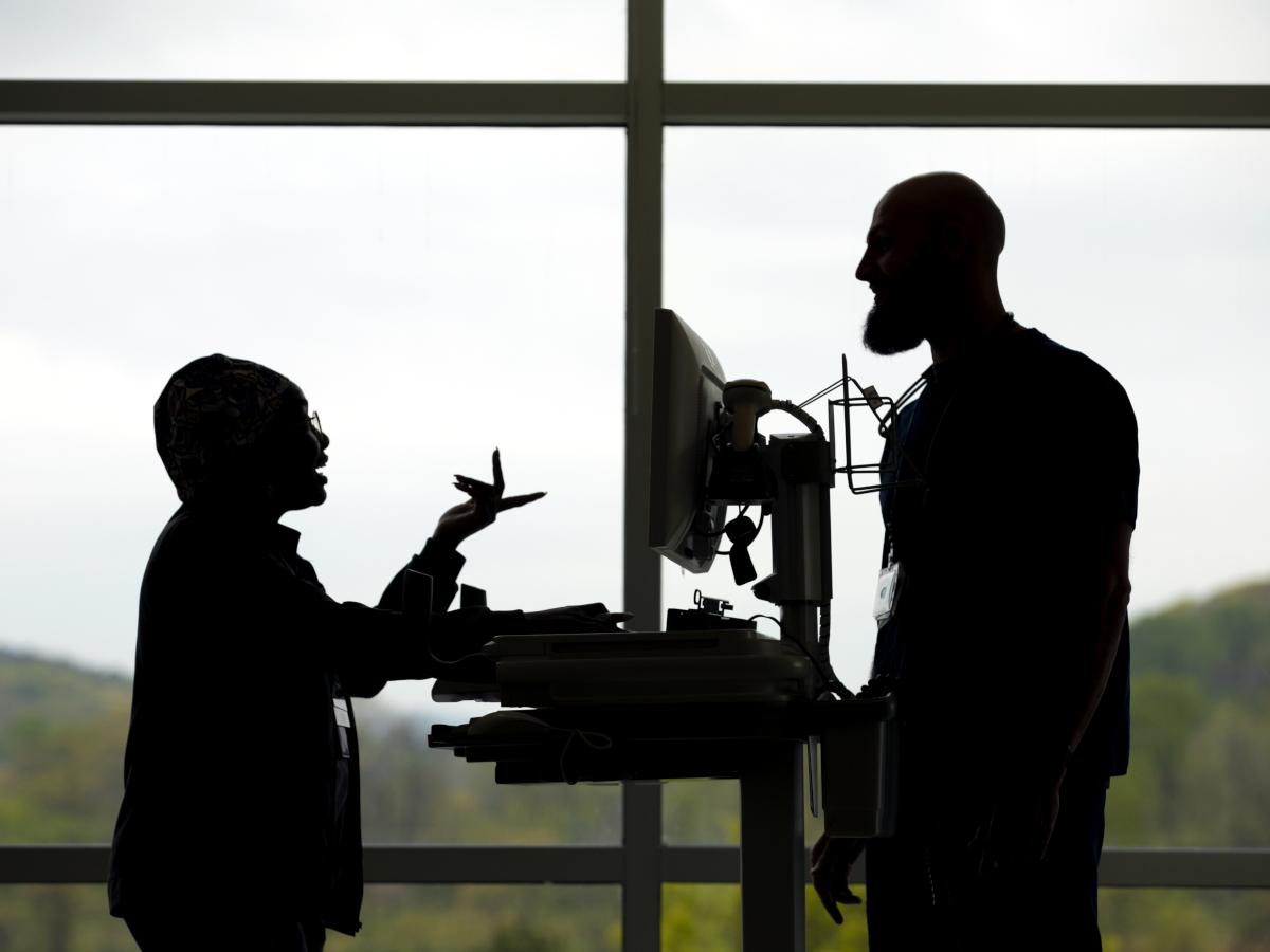 A silhouette of two employees standing and talking in front of a window at Penn State Health St. Joseph Medical Center. A computer on a cart is between the two of them.