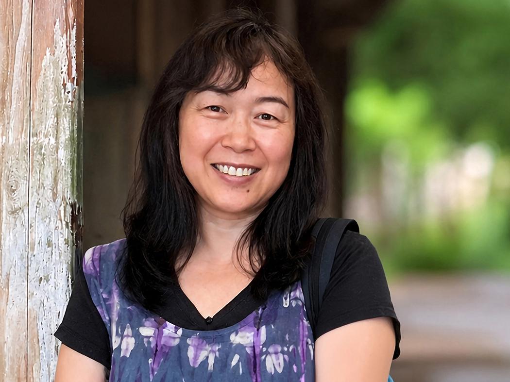 Portrait of a person with shoulder-length dark hair, wearing a purple patterned top over a black shirt, standing beside a weathered wooden post with a blurred outdoor background.