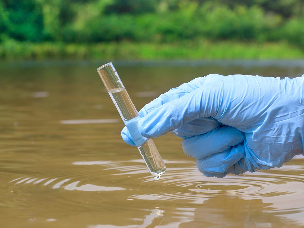 a gloved hand holds a beaker of water above a body of water