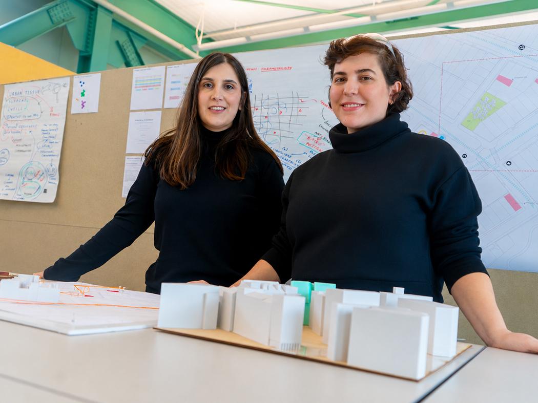Two people stand behind a tabletop architectural model in a design studio, with planning sketches, diagrams, and a site map displayed on boards in the background.