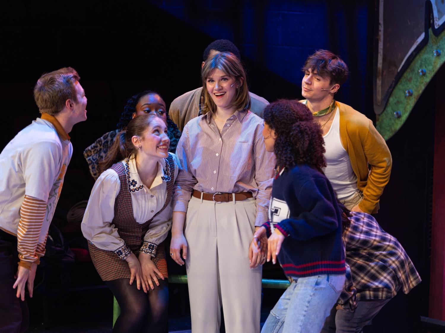 A woman in blouse and slacks surrounded by actors/singers