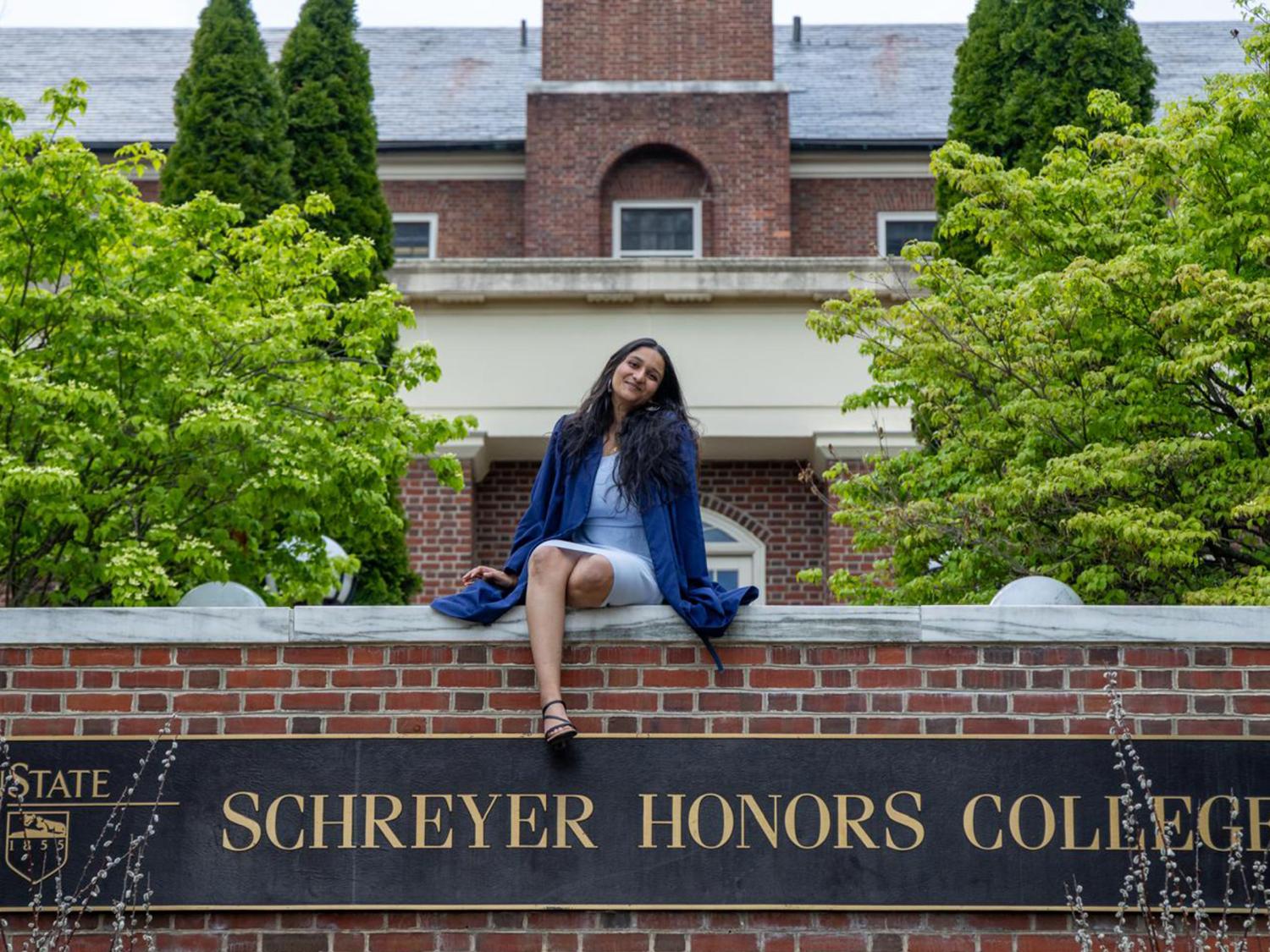 A person in a graduation gown sits on a brick wall with a large plaque reading "Schreyer Honors College" 
