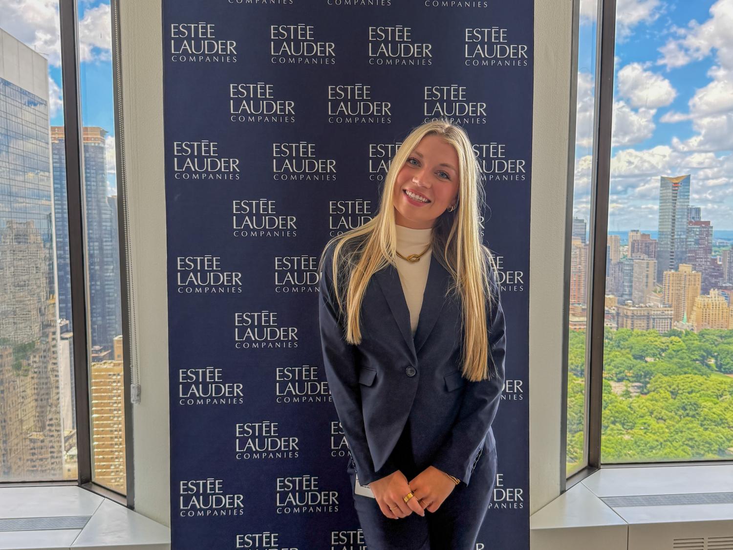 Karli Brenneman wears a black suite while standing in front of a banner that reads “Estée Lauder Companies” with the New York City skyline out the window next to her.