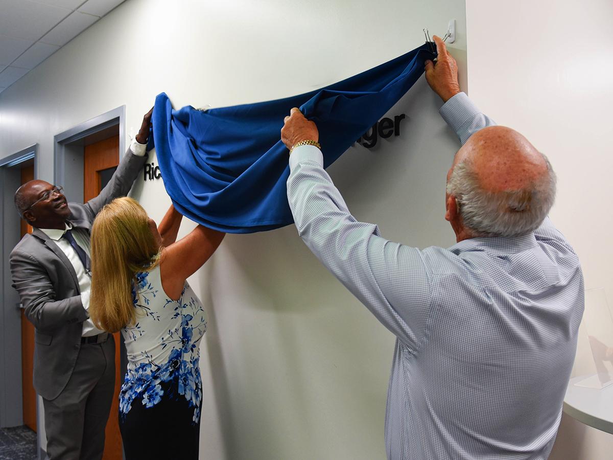 Ron Darbeau, Jeane Singer, and Richard Singer unveil a plaque in a hallway.