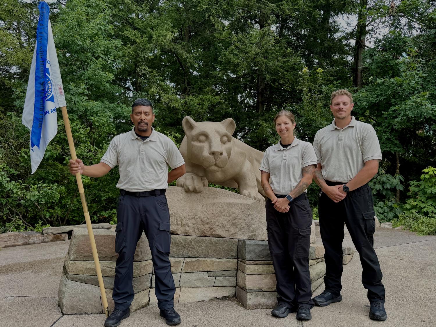 three people with a blue and white flag at the nittany lion statue