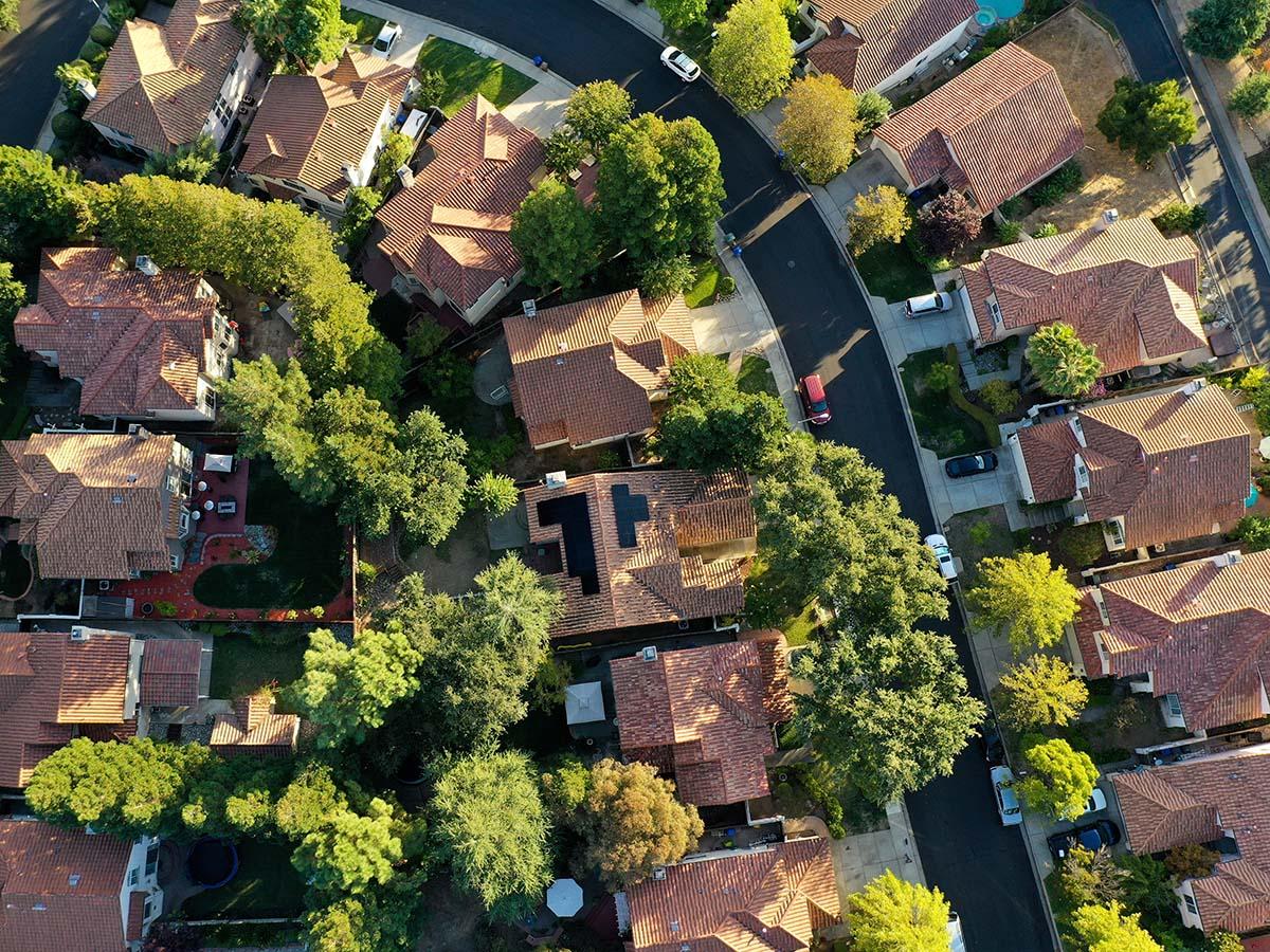 Aerial view of a suburban neighborhood with red-tiled roofs, surrounded by green trees and neatly paved streets, showing several homes and yards.