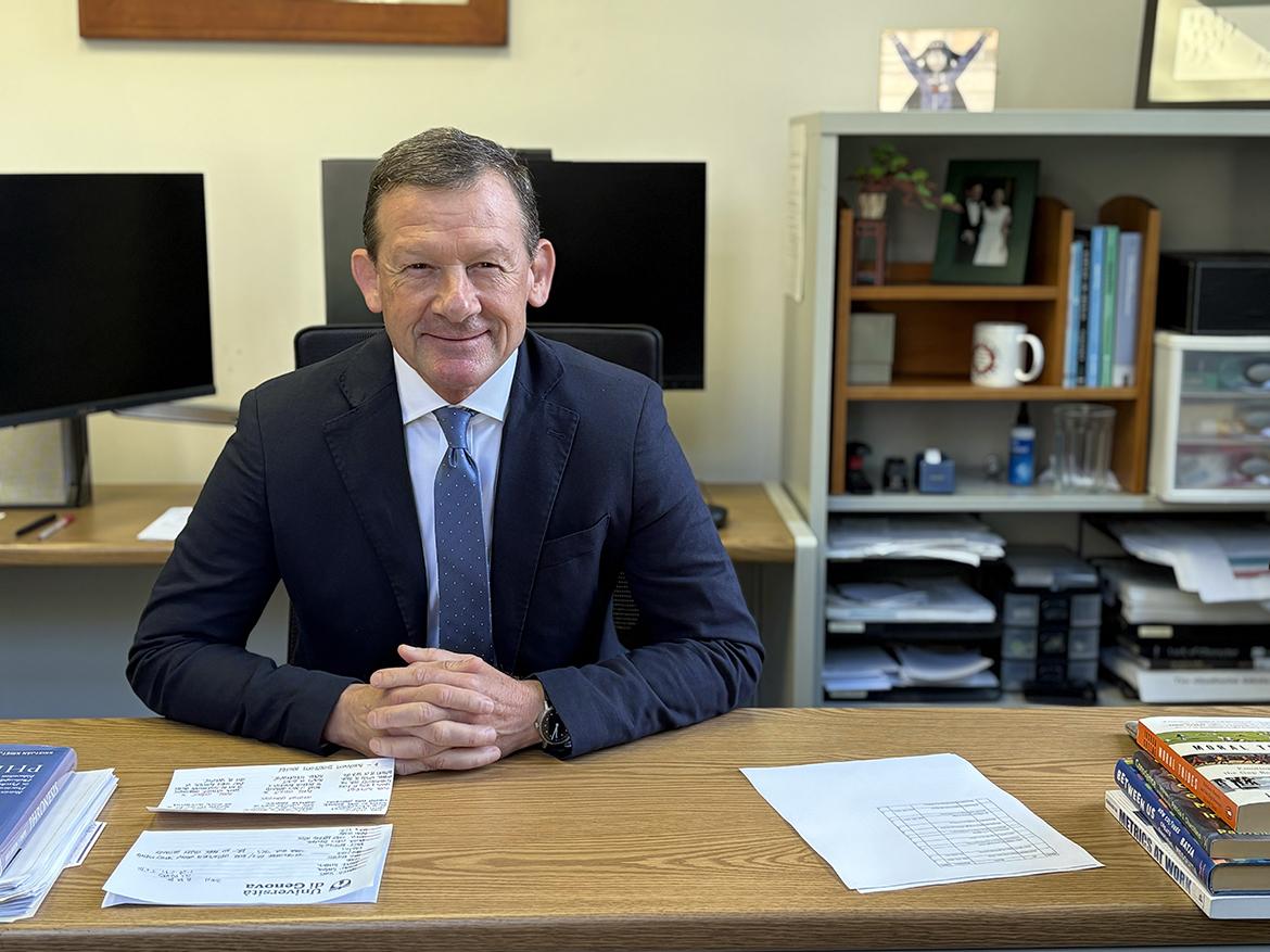 A man in a suit and tie sits at a desk and smiles at the camera.