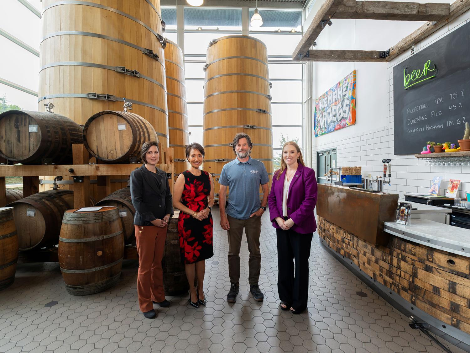 Four people stand in a large room with wooden barrel kegs stacked on one side