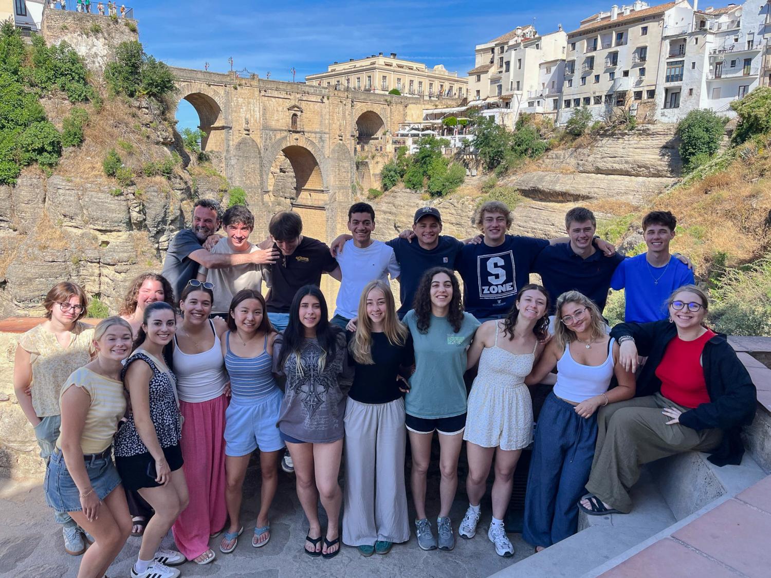 A group of Penn State students standing together, posed against a backdrop of a picturesque, historical hillside with colorful buildings and a stone bridge.