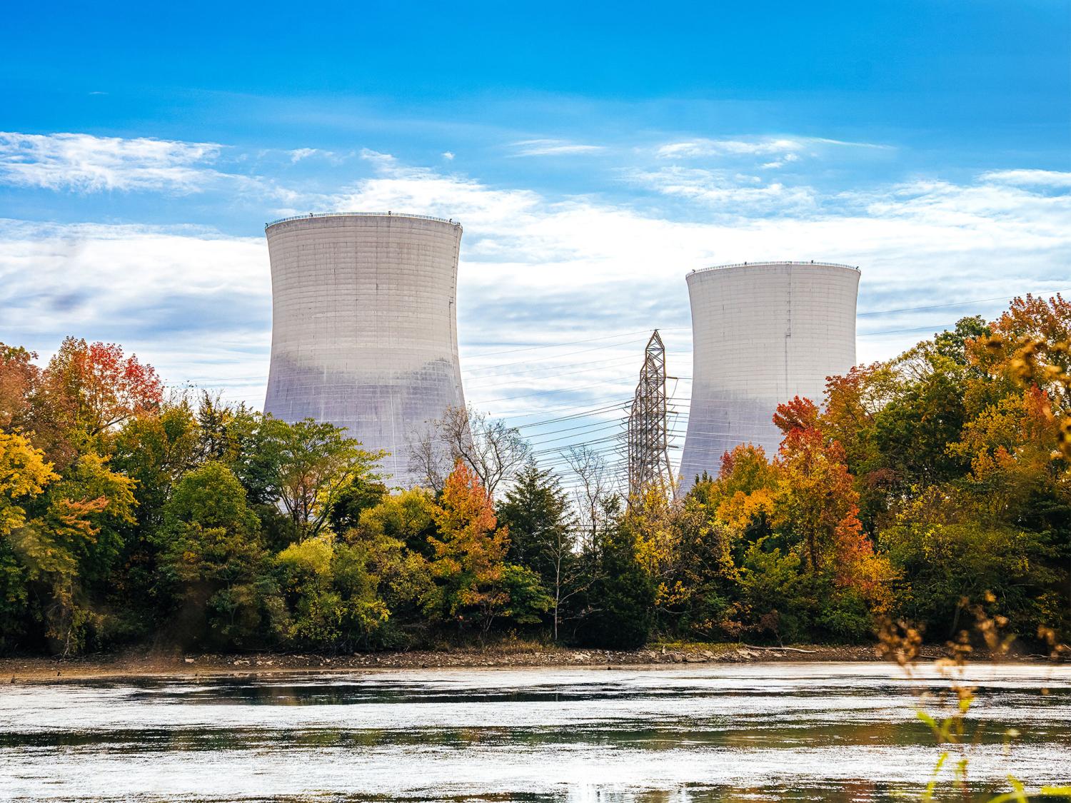 A view over a lake shows two large cooling towers of a nuclear power plant overlooking some trees on a sunny day