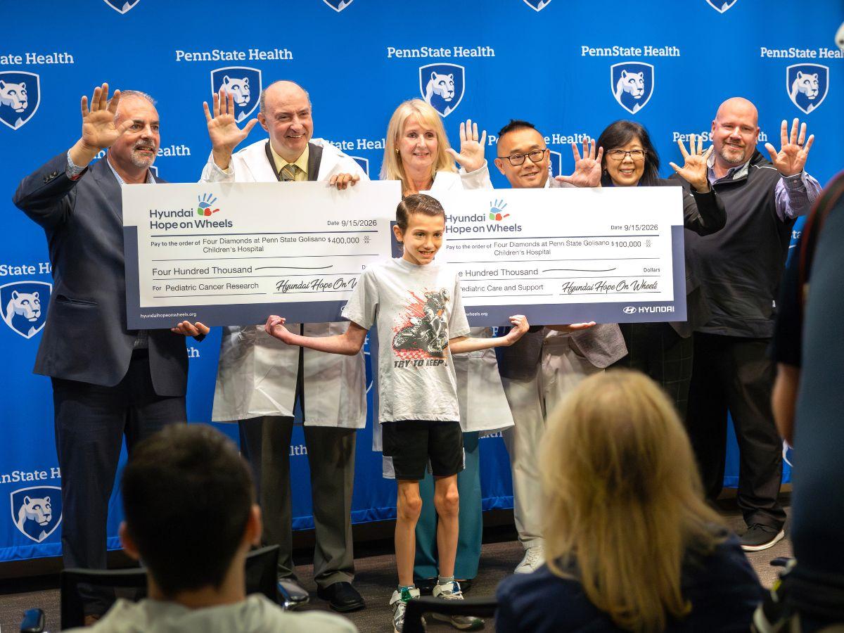 Group of Penn State Health physicians and staff hold oversized Hyundai Hope on Wheels checks totaling $500,000 for pediatric cancer research and care, with a child standing in front of the group.