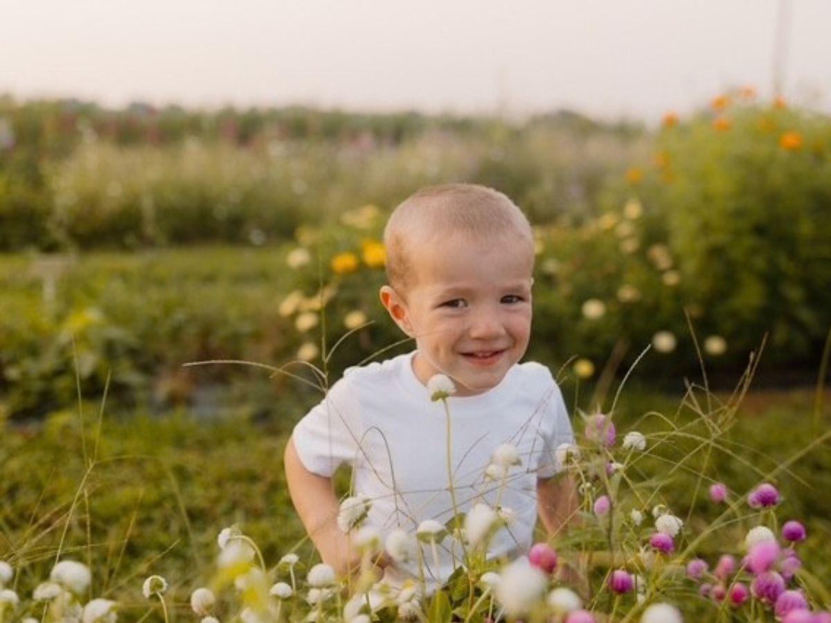 A young child wearing a white T-shirt sits among blooming wildflowers in an outdoor garden or flower field.