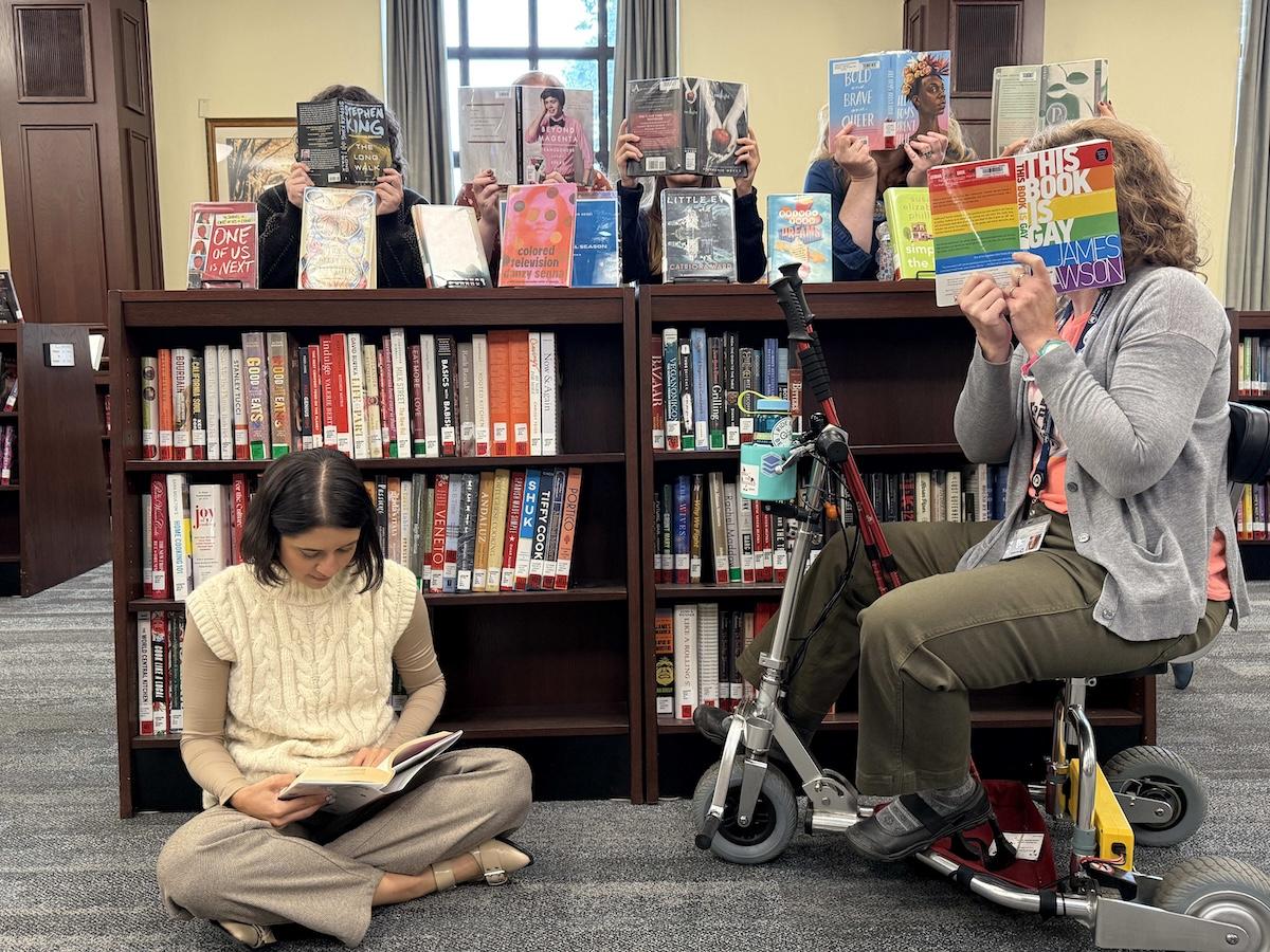 individuals sitting and standing with banned books open in front of their faces, around a bookshelf