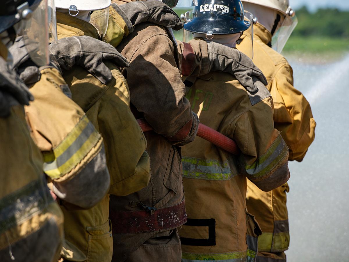 Firefighters in protective gear work together near a road, carrying a hose.