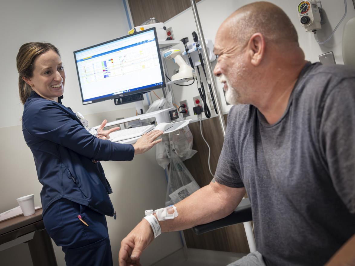 A patient sits in a chair in a clinical room, looking over at a nurse who stands at a computer terminal. She looks back, smiling.