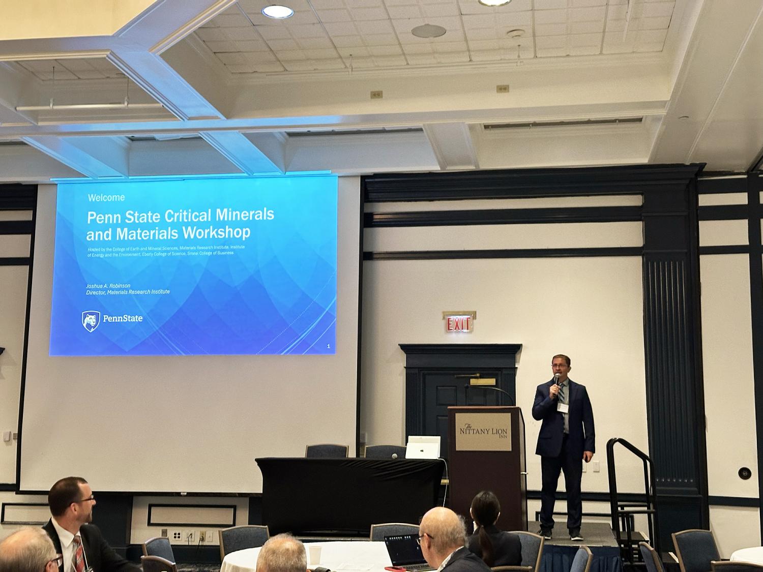 A man in a suit speaking into a microphone to the right of a blue projected slide that reads "Penn State Critical Minerals and Materials Workshop"