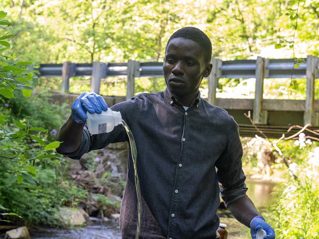 Person stands in a stream, pouring out a sample of water. 