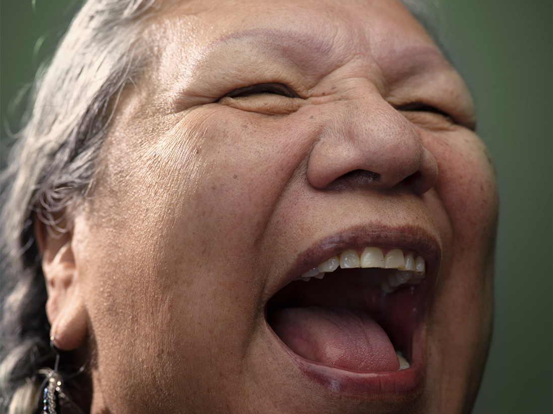 Photo portrait of Lakota Harden, Native community leader and elder, laughing broadly, open-mouthed. Her face, which makes up most of the image, is framed by gray hair in braids, dangling blue earrings, and a dark green background.