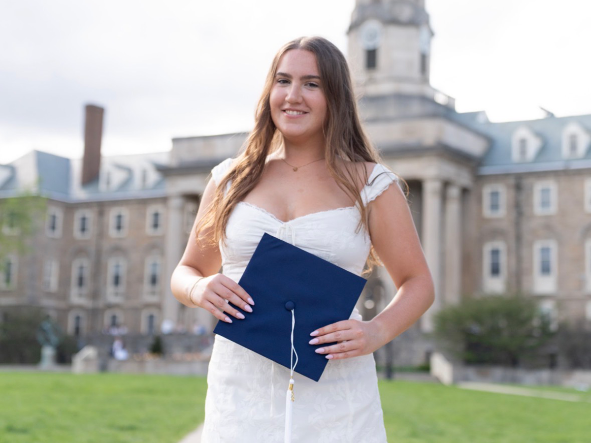 Marsella smiles while holding a dark blue graduation cap in front of Old Main. She is wearing a white dress.