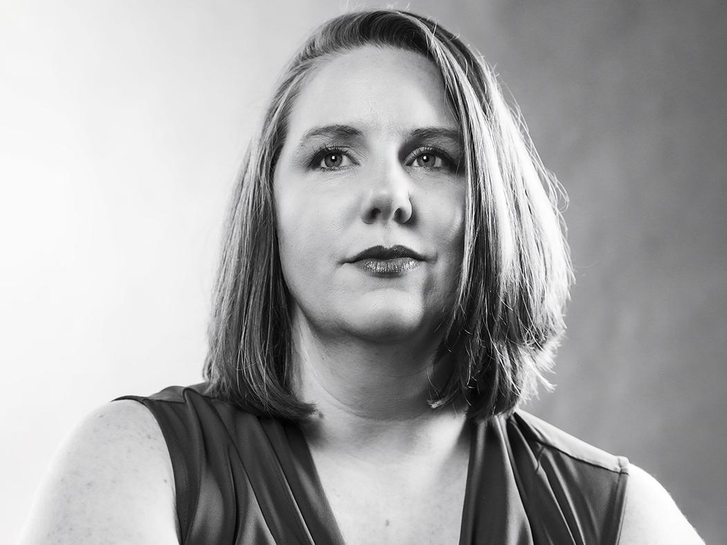 Black-and-white head-and-shoulders portrait of a person with shoulder-length hair, facing slightly to the side, wearing a sleeveless top against a softly blurred background.