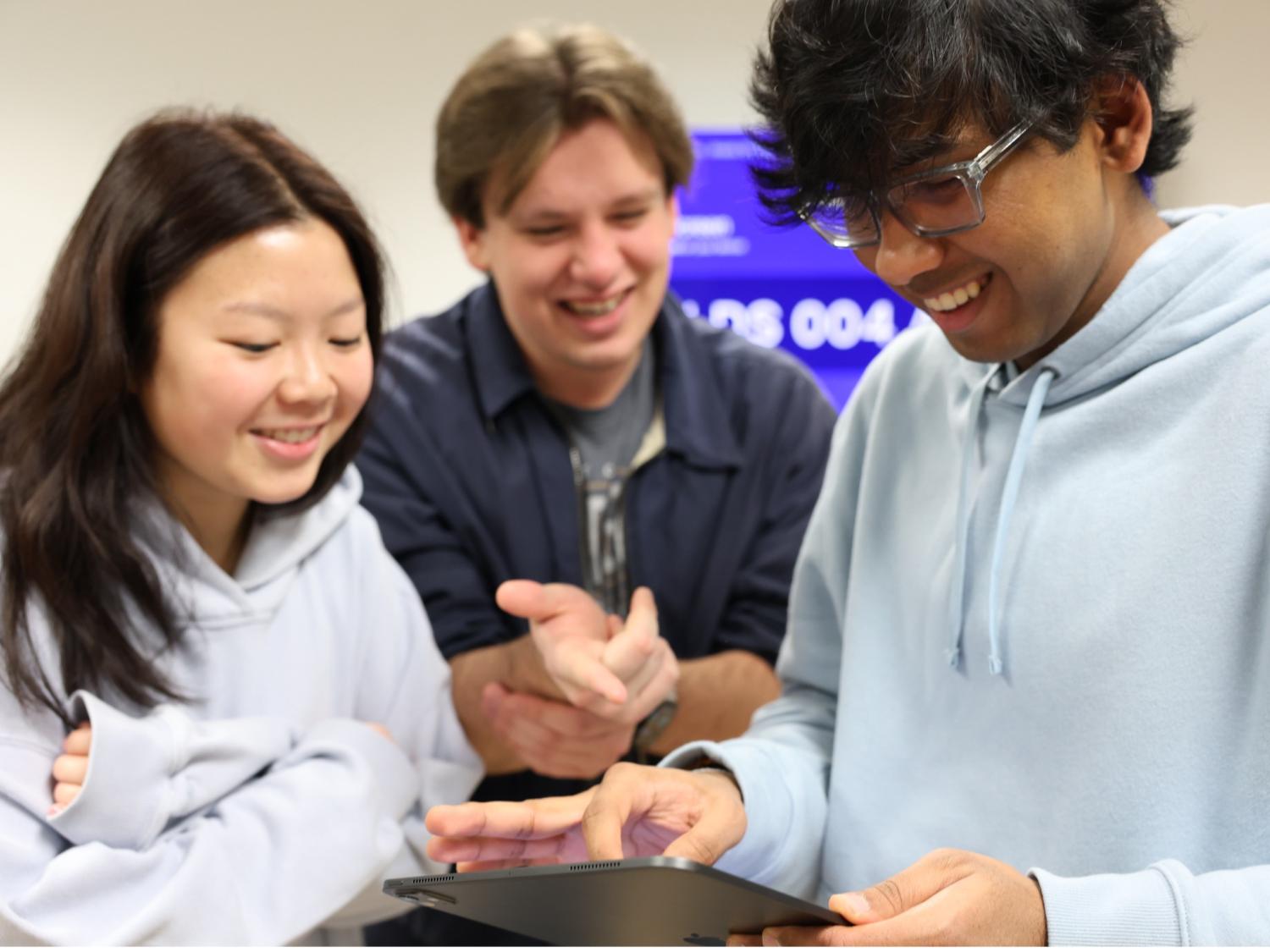 Three smiling students gather around an iPad. One holds the tablet and taps the screen while the other two look on. A purple presentation screen is blurred in the background.