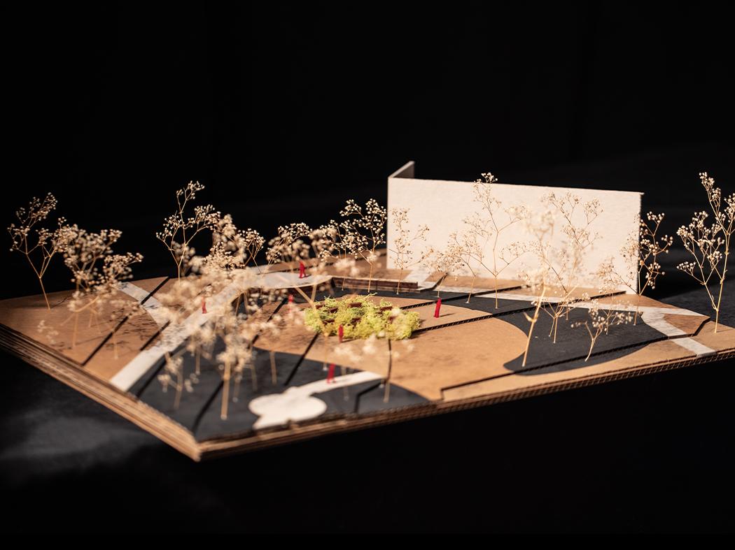 Architectural site model showing a proposed building, pathways, trees, and landscaped gathering spaces arranged on a cardboard base against a dark background.