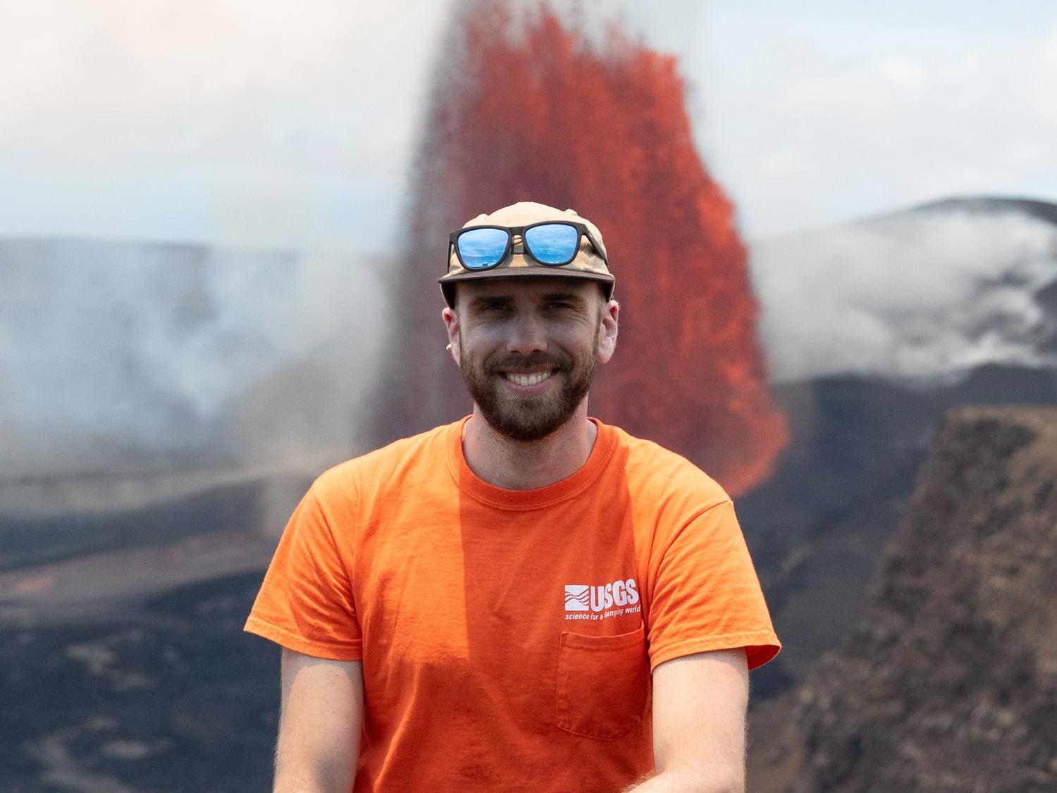 A bearded man wearing an orange USGS shirt and sunglasses balanced on the brim of a baseball cap standing in front of a lava spurt of an active volcano