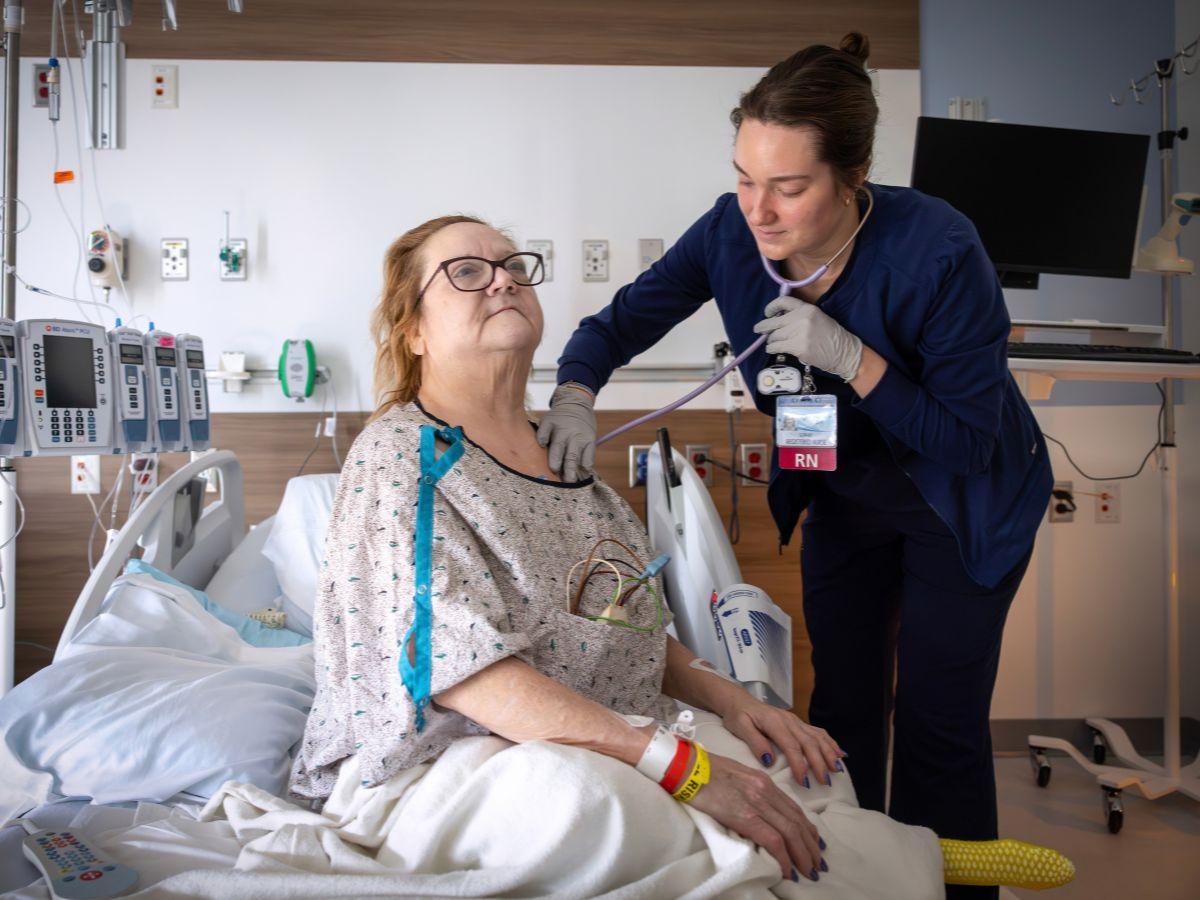 A nurse examines a hospitalized patient using a stethoscope while the patient sits upright in a hospital bed.