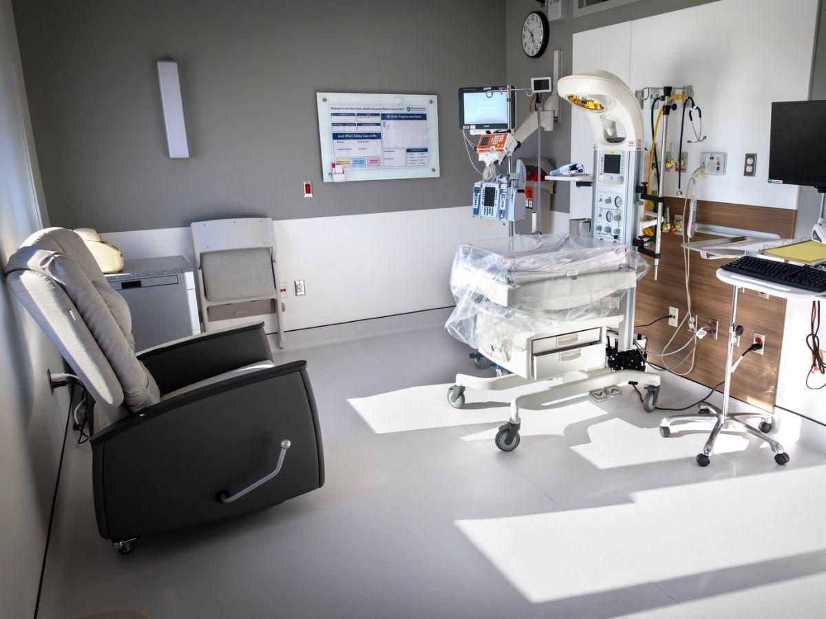 A private neonatal intensive care unit (NICU) room at Penn State Health Lancaster Medical Center featuring an infant radiant warmer with monitoring equipment, IV pumps, and respiratory support equipment beside a computer workstation. A cushioned recliner and chair provide space for parents, while large windows fill the room with natural light.