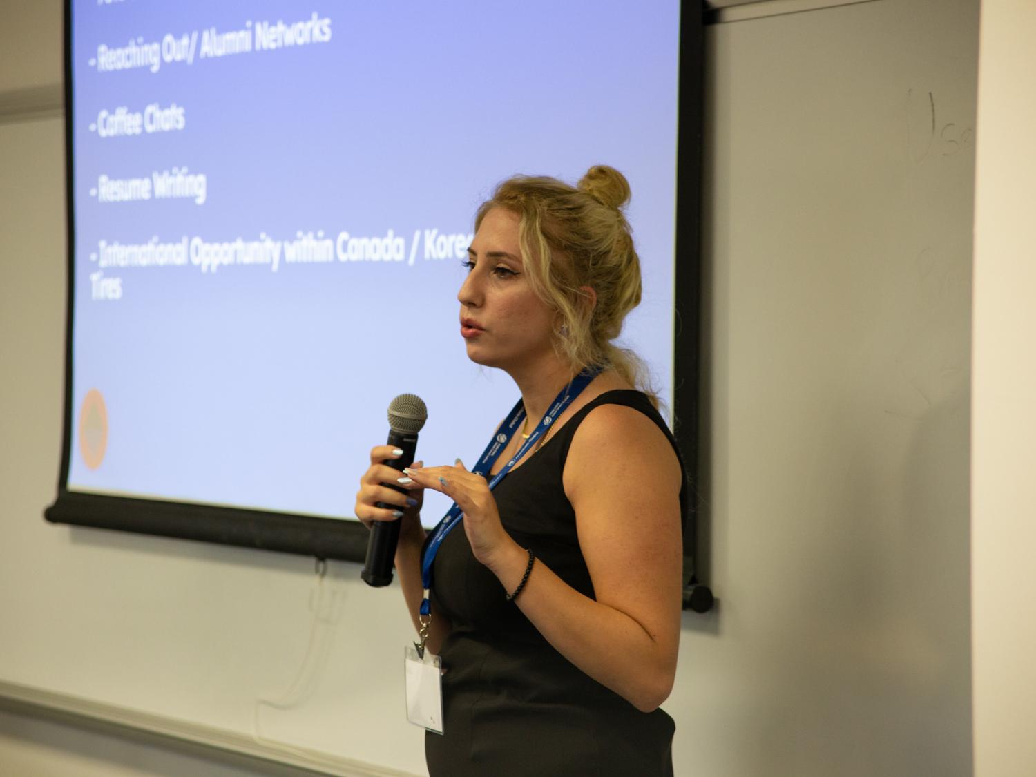 A woman speaks into a microphone in front of a screen with a slideshow.