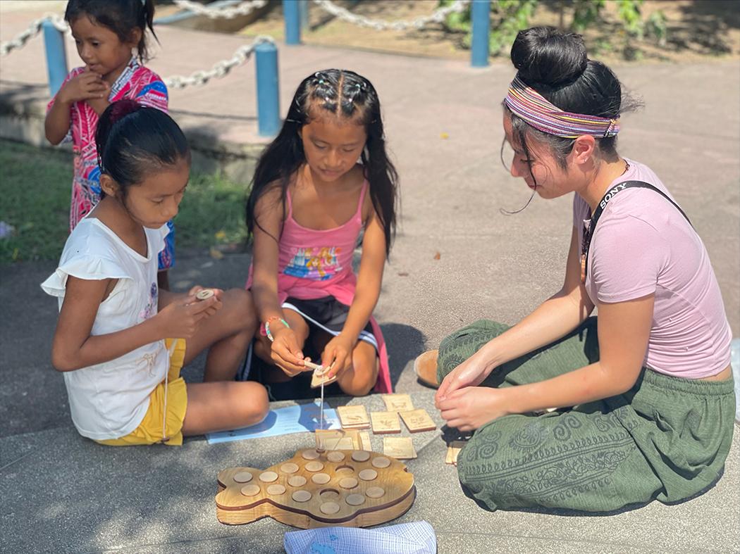 Three children and an adult sit outdoors around a wooden learning game. Two children handle game pieces while another watches; the adult sits nearby, observing. The game board and pieces are spread on the pavement.