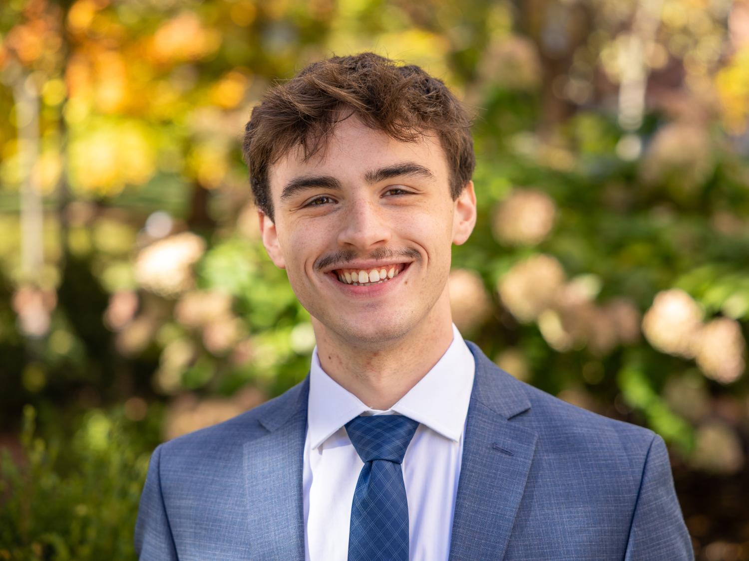 Dawson Glogau stands outdoors near Old Botany, wearing a blue-grey jacket, white collared shirt and a blue tie.
