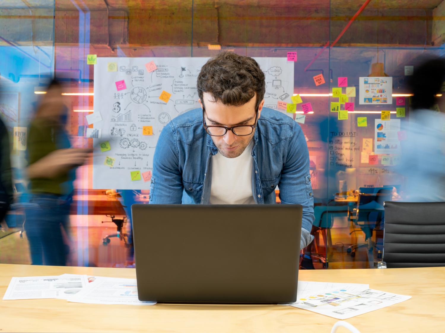 Young man with glasses examines a laptop in a bustling office environment