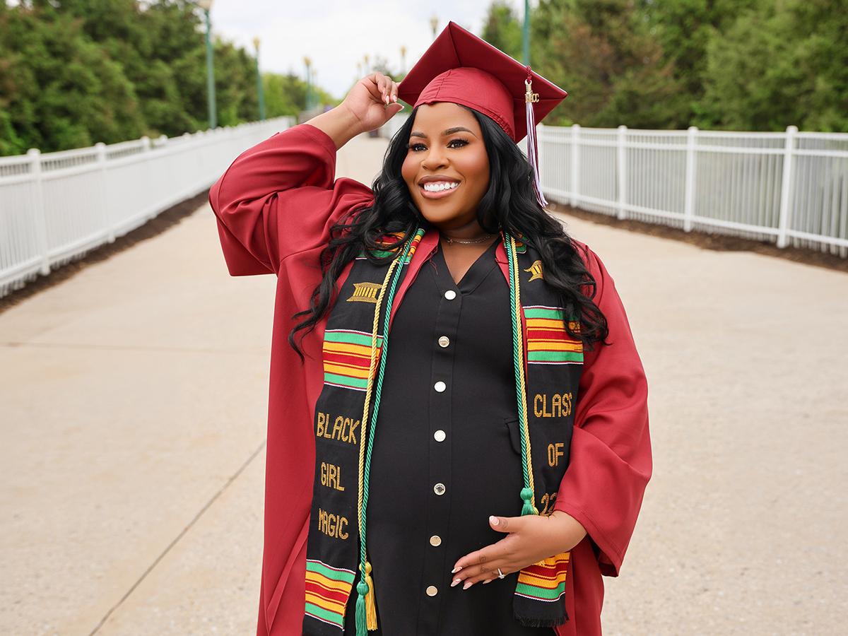 A smiling pregnant woman poses outdoors in a red graduation cap and gown, wearing a black dress and a colorful stole reading “Black Girl Magic” as she cradles her belly.