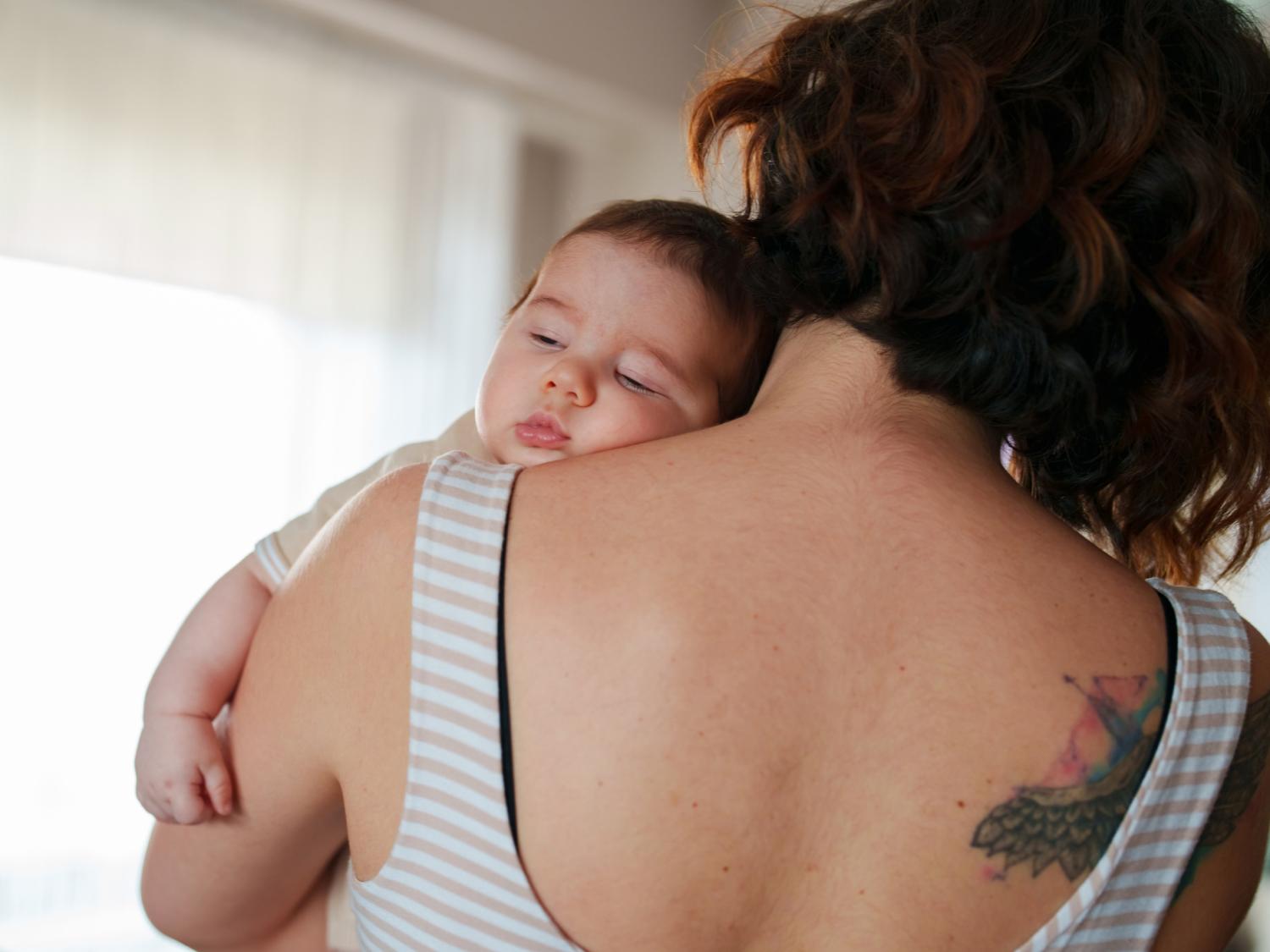 A person in a tank top is facing away from the camera while a sleepy-looking baby rests on their shoulder, facing the camera.