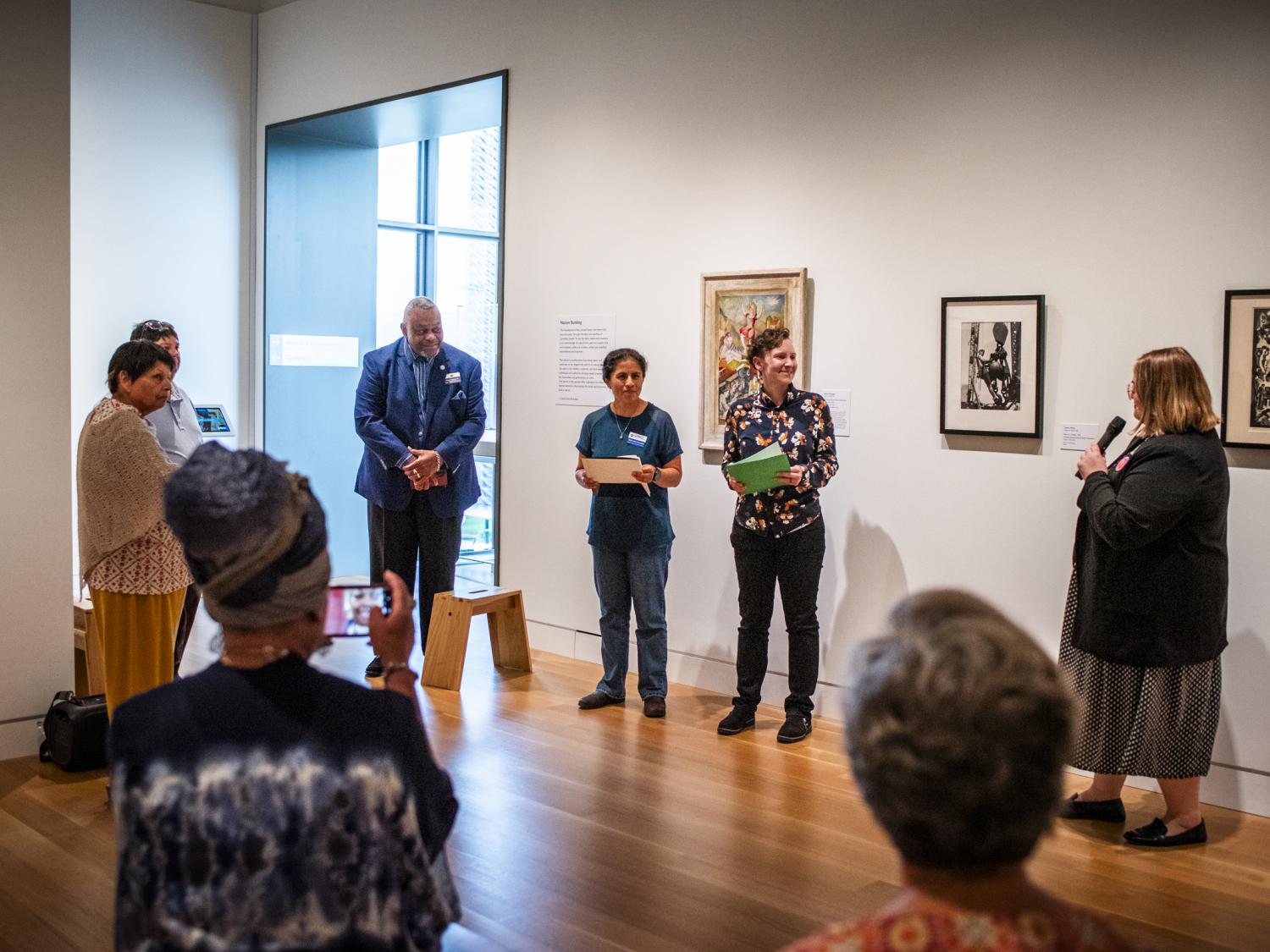 A group of people standing in an art museum. The person at right holds a mic and is introducing the three people to her left.