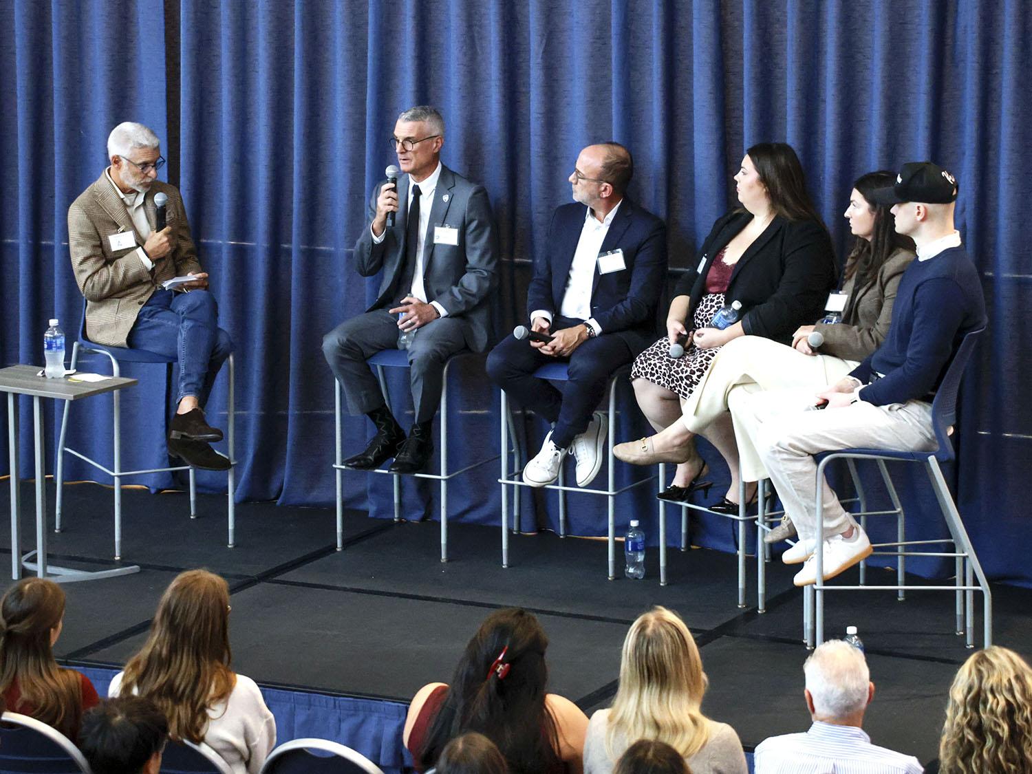 Five panelists sit on raised stools on a platform answering questions from a moderator