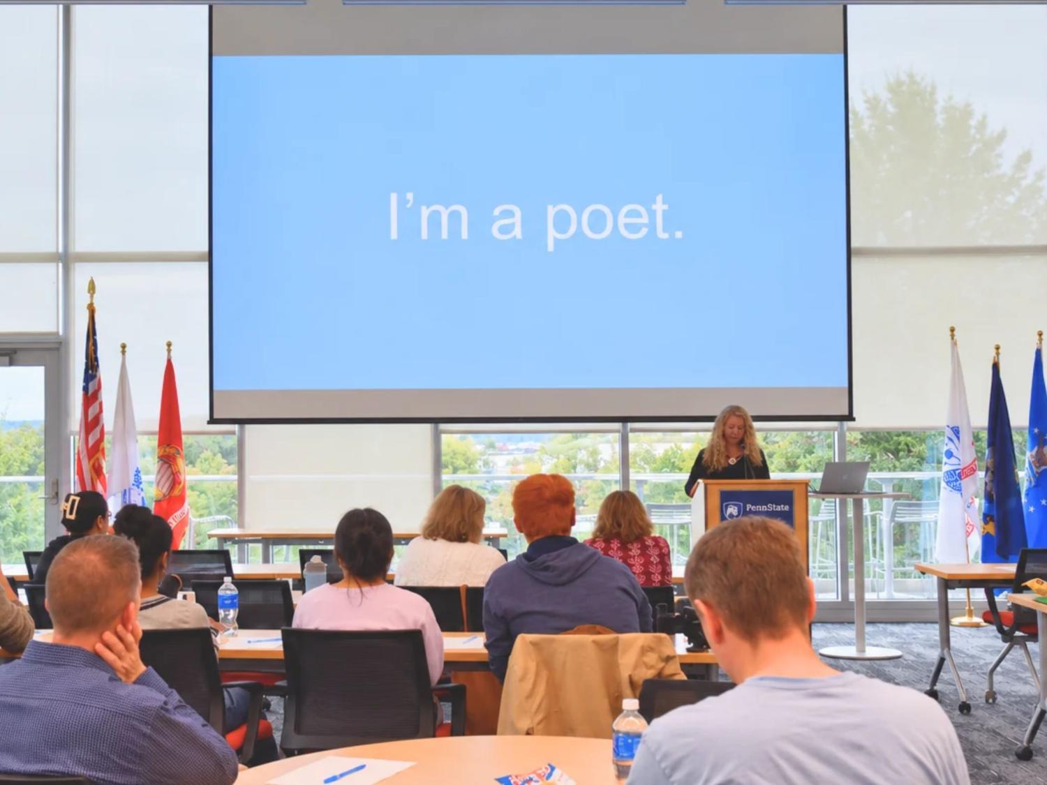 Audience members listen as Penn State Laureate Erin Murphy speaks from a podium beneath a projected slide reading, “I’m a poet.”