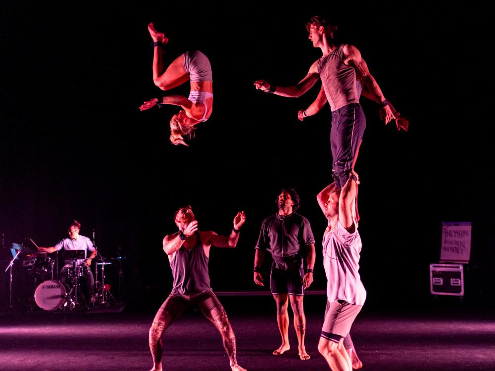 A drummer performs while acrobats do mid-air flips and handstands.