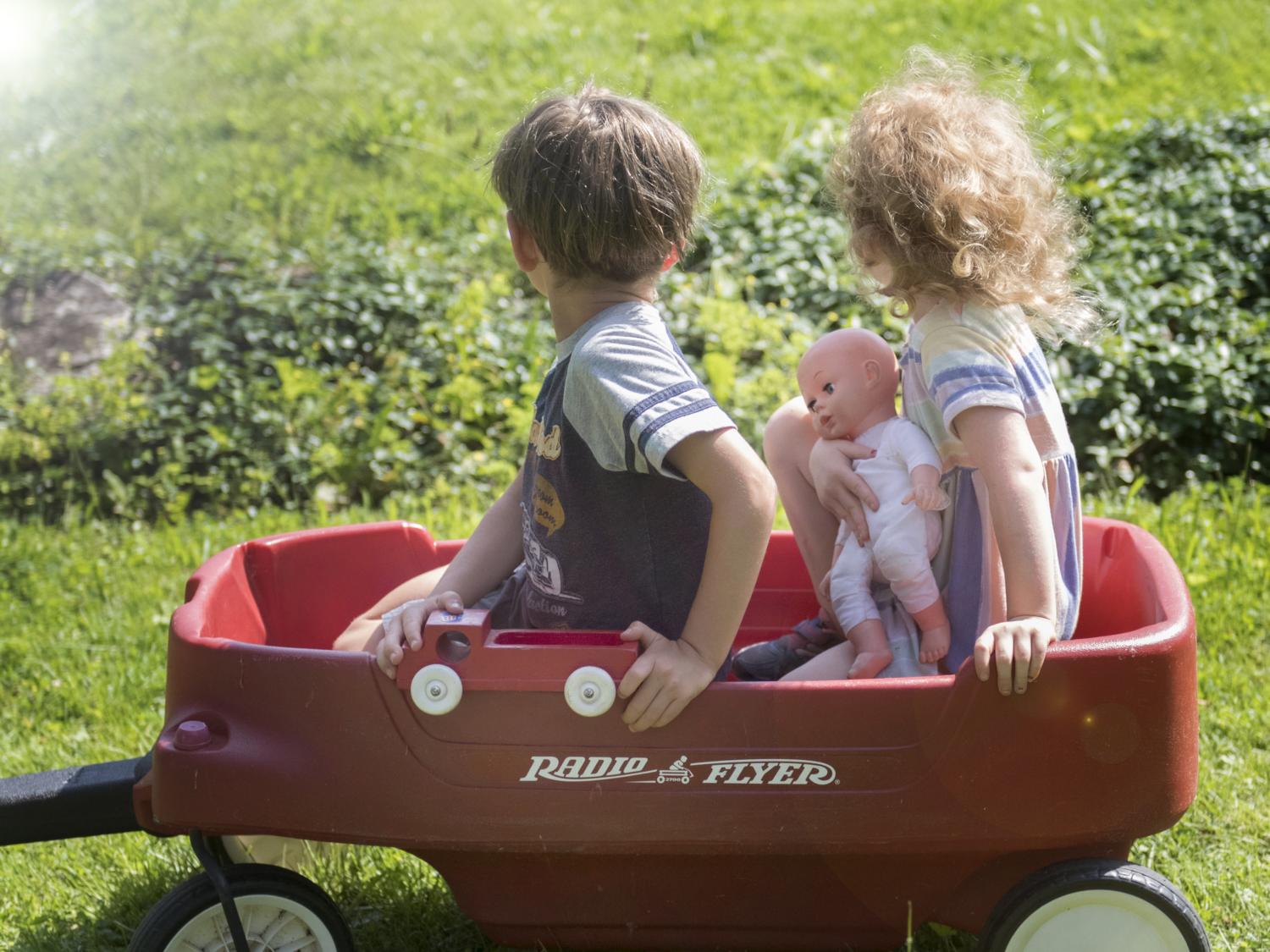 A little boy and little girl sitting in a red wagon.  The boy has a red truck and the girl is holding a doll.