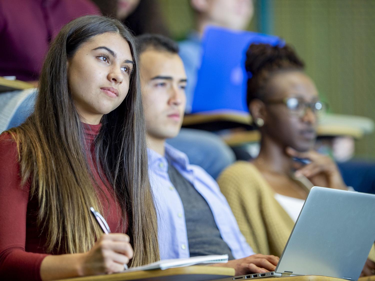 Two young women and a young man sit in a lecture style hall while taking class notes.