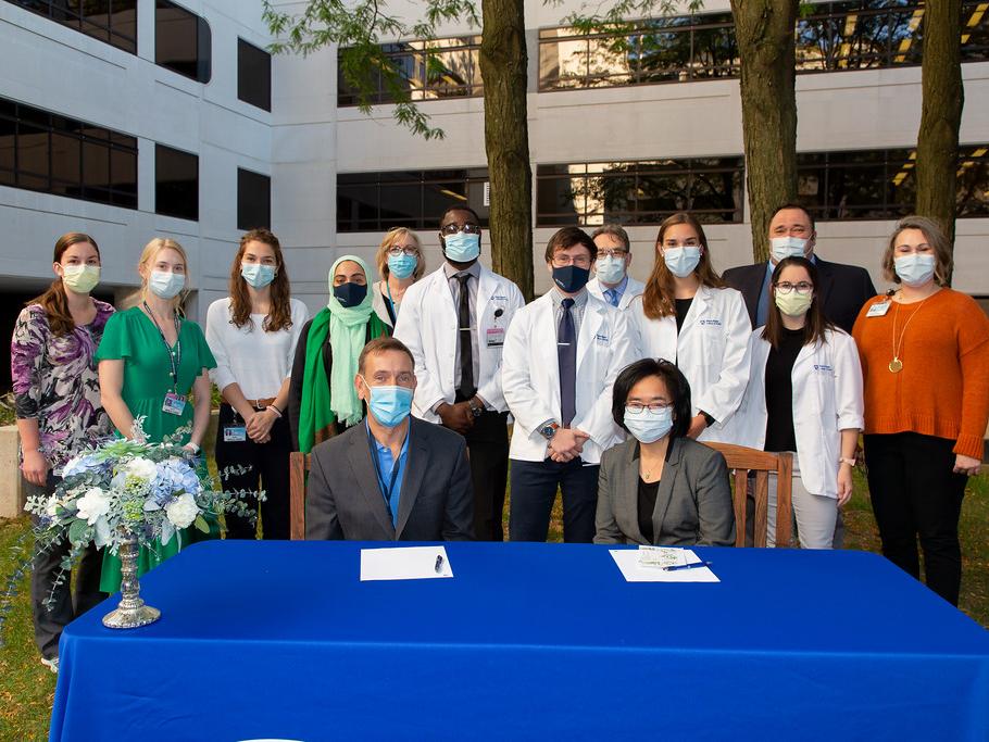 A group of medical students stand behind a table with the Dean of the College of Medicine and another woman seated at a table in front of them. Everyone is outdoors and wearing masks.
