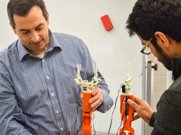 professor in collared shirt holds a robot arm, while student to the right of professor holds another robot arm