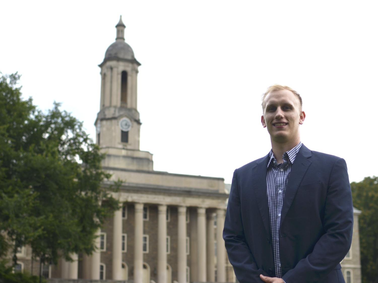 A man in his 20s in a blue blazer, blue and white checked shirt, stands in front of Old Main bell tower at Penn State University Campus. 